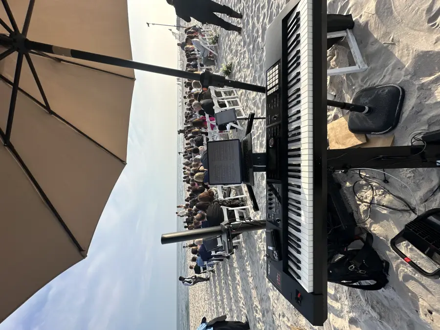 Isaac's keyboard set up under an umbrella at a beach wedding ceremony, guests facing the ocean