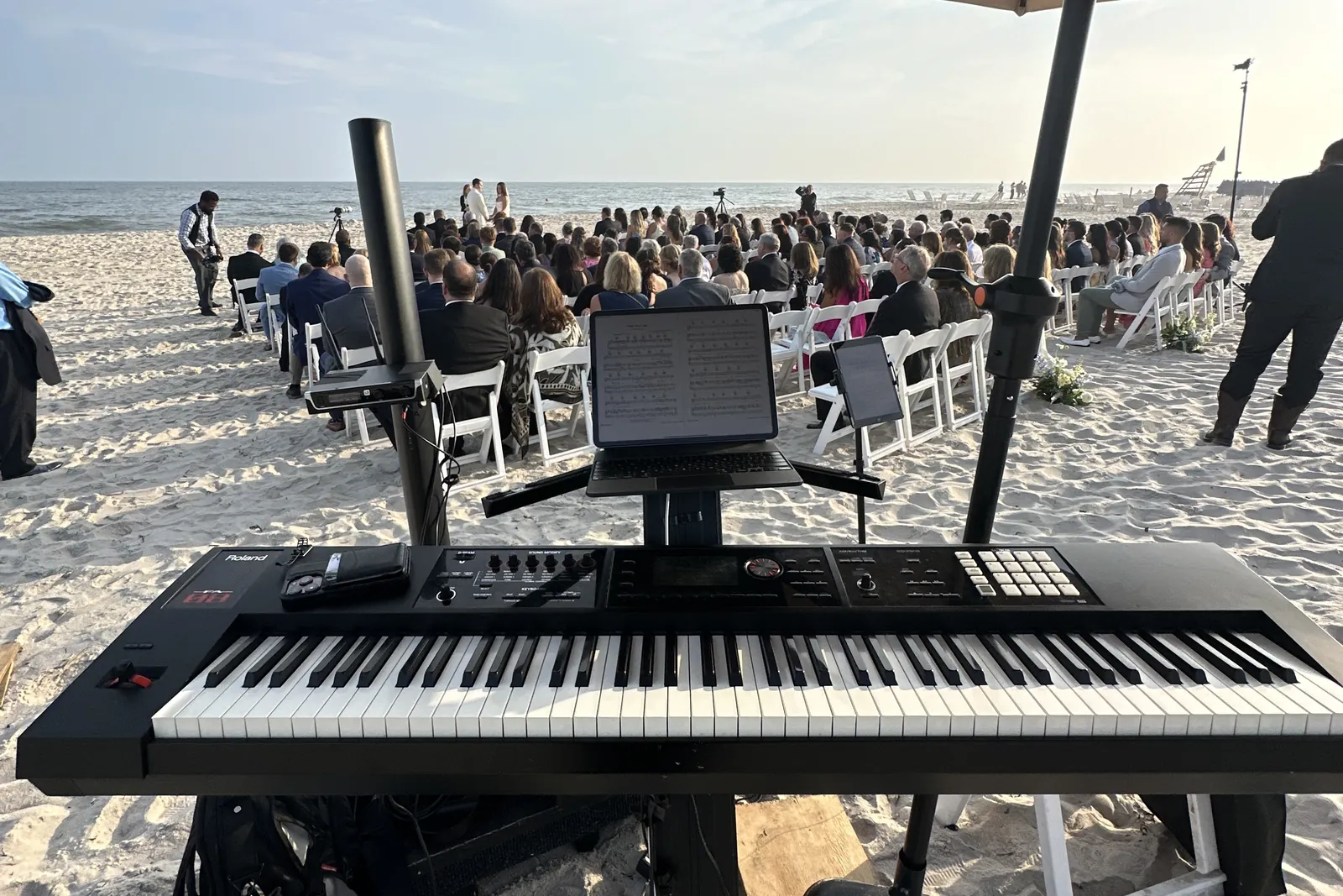 Keyboard under an umbrella at a beach ceremony, guests facing the ocean