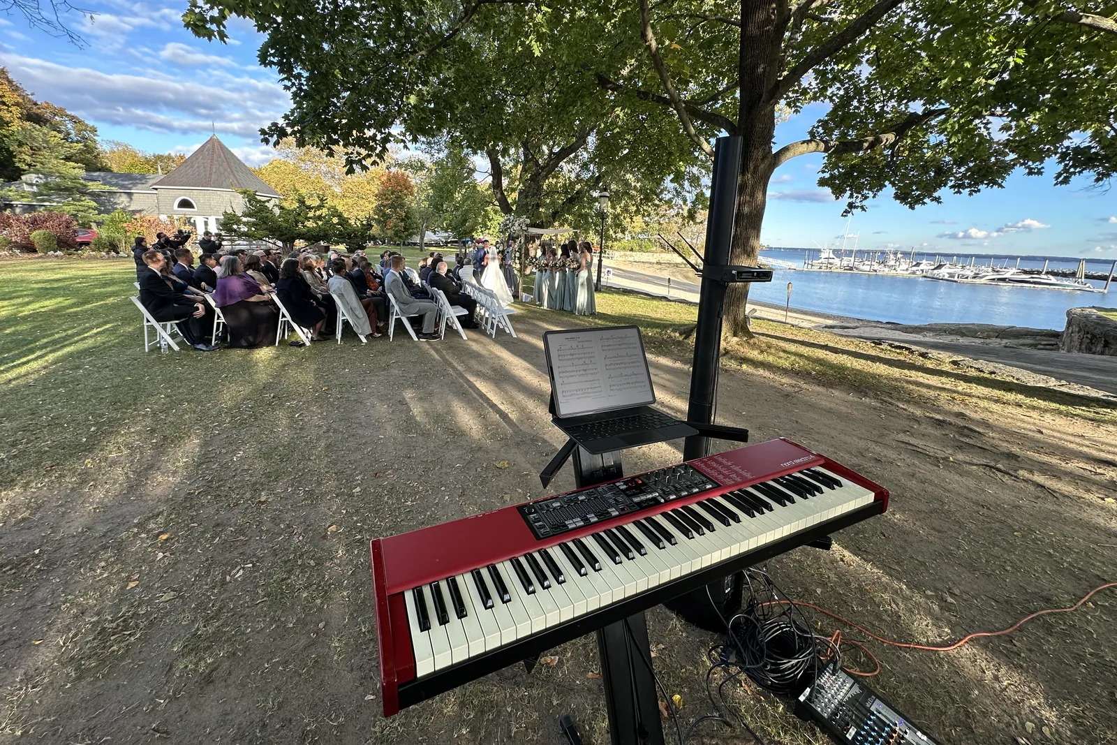 Red stage piano on a lawn under a tree, guests seated by the water